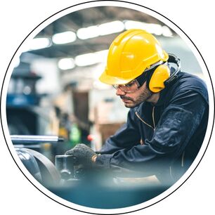Das Bild zeigt eine Person in einem industriellen Umfeld. Sie trägt einen gelben Schutzhelm und gelbe Gehörschutzkopfhörer und arbeitet an einer Maschine. Ist das so in Ordnung?