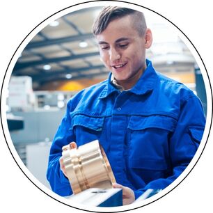 Eine Person in blauer Arbeitskleidung hält eine große, zylindrische, metallische Komponente in einer Werkstatt oder industriellen Umgebung. Im Hintergrund sind verschiedene Maschinen und Geräte zu sehen, was auf ein Fertigungs- oder Ingenieurumfeld hinwei