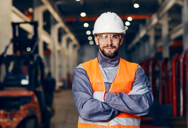 Die Person auf dem Bild trägt einen weißen Schutzhelm, eine Schutzbrille und eine orangefarbene Sicherheitsweste. Sie steht mit verschränkten Armen in einem industriellen Umfeld, das auf Sicherheit und Schutzmaßnahmen hinweist.